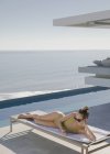 Mulher banhos de sol, mensagens de texto com telefone inteligente na cadeira lounge no pátio de luxo ensolarado com vista para o mar — Fotografia de Stock