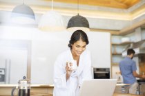 Mujer en albornoz beber café y el uso de ordenador portátil en la cocina - foto de stock