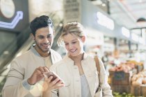 Усміхнена молода пара використовує мобільний телефон на ринку продуктових магазинів — стокове фото