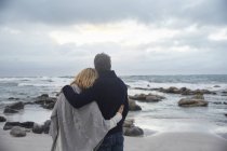Спокойная ласковая пара, обнимающаяся на зимнем пляже и смотрящая на океан — стоковое фото