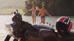 Молода пара біжить на причалі біля озера до мотоцикла — стокове фото