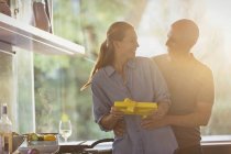 Smiling husband surprising wife with gift in sunny kitchen — Stock Photo