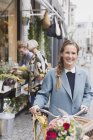 Портрет улыбающейся женщины, идущей на велосипеде с цветами в корзине перед магазином — стоковое фото