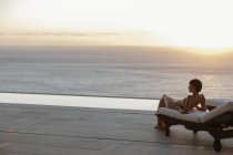 Femme en robe posée sur une chaise longue sur un patio surplombant l'océan au coucher du soleil — Photo de stock