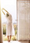 Woman in bathrobe stretching with arms overhead at sunny patio doorway — Stock Photo