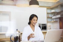 Mujer risueña en albornoz bebiendo café y usando laptop - foto de stock
