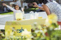 Mother photographing daughter at lemonade stand — Stock Photo