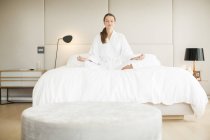 Serene woman in bathrobe meditating in lotus position on bed — Stock Photo
