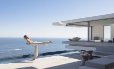 Frau praktiziert Yoga-Krieger 3 Pose auf sonnigen modernen, luxuriösen Haus Vitrine Außenterrasse mit Meerblick — Stockfoto