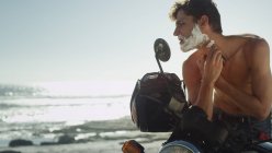 Young man on motorcycle shaving beard near ocean — Stock Photo