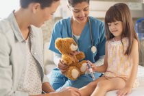 Жіноча медсестра з плюшевим ведмедем дивиться дівчину-пацієнтку за допомогою інсулінової ручки в лікарняній кімнаті — стокове фото