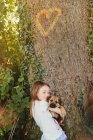 Chica sosteniendo cachorro perro debajo del árbol con forma de corazón - foto de stock