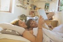 Улыбающаяся молодая пара, лежащая в постели с помощью цифрового планшета — стоковое фото