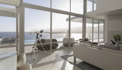 Sunny, casa de luxo moderno tranquilo vitrine interior sala de estar com telescópio e vista para o mar — Fotografia de Stock