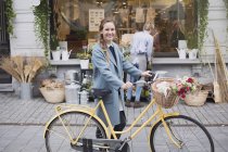 Портрет улыбающейся женщины, идущей на велосипеде с цветами в корзине перед магазином — стоковое фото