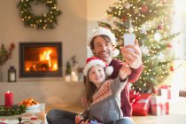 Padre e hija en los sombreros de Santa tomar selfie con el teléfono de la cámara en la sala de estar de Navidad - foto de stock