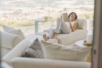 Усміхнена жінка читає книгу, розслабляючись у вітальні — стокове фото