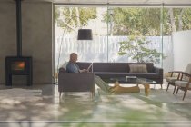 Homem maduro relaxante, usando tablet digital no sofá na sala de estar com lareira — Fotografia de Stock