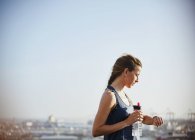 Corredor feminino com garrafa de água verificando relógio inteligente rastreador de fitness sob céu bleu ensolarado — Fotografia de Stock