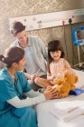 Female nurse and girl patient using digital thermometer on teddy bear in hospital room — Stock Photo