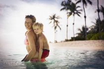 Ritratto sorridente madre piggybacking figlio nell'oceano tropicale — Foto stock