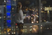 Пенсионерка с цифровым планшетом, работающая допоздна, выглядывающая ночью из окна городского офиса — стоковое фото