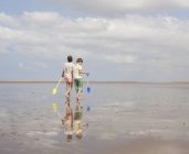 Hermano y hermana caminando con palas en arena mojada en la soleada playa de verano - foto de stock