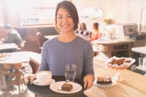 Portrait smiling waitress carrying tray with cappuccino, brownie and water in cafe — Stock Photo