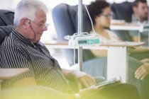 Paciente mayor que recibe tratamiento mientras está sentado y leyendo un libro en la sala del hospital - foto de stock