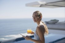 Портретна жінка використовує цифровий планшет і п'є апельсиновий сік на сонячному розкішному дворику з видом на океан — стокове фото