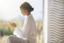 Mujer en albornoz mensajes de texto con teléfono celular en la puerta soleada - foto de stock