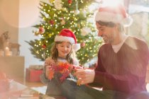 Father and daughter wearing Santa hats and holding paper snowflakes near Christmas tree — Stock Photo