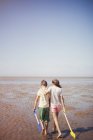 Brother and sister with shovels hugging and walking in wet sand on sunny summer beach below blue sky — Stock Photo