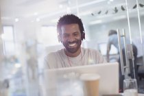 Усміхнений, впевнений креативний бізнесмен з навушниками, використовуючи ноутбук в офісі — стокове фото
