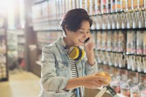 Smiling young woman with headphones talking on cell phone grocery shopping in market — Stock Photo