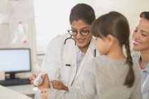 Pediatra femenina mostrando termómetro digital a niña paciente y madre en sala de examen - foto de stock