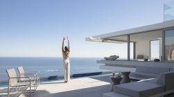 Donna che pratica yoga montagna posa sul soleggiato moderno, casa di lusso vetrina patio esterno con vista sull'oceano — Foto stock