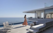 Donna in abito camminare su soleggiato, moderno, casa di lusso vetrina patio esterno con vista sull'oceano — Foto stock