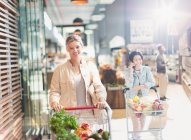 Portrait smiling young woman with shopping cart in grocery store market — Stock Photo