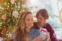 Чоловік дивиться, як дружина відкриває різдвяний подарунок перед ялинкою — стокове фото