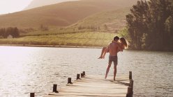 Young man carrying woman on sunny lakeside dock — Stock Photo