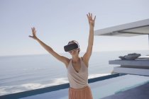 Енергетична жінка використовує окуляри симулятора віртуальної реальності на сучасному, розкішному домашньому вітрині зовнішнього дворику з видом на океан — стокове фото