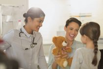 Mujer sonriente pediatra y madre mostrando oso de peluche a niña paciente en sala de examen - foto de stock