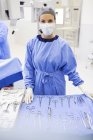 Retrato de enfermera quirúrgica de pie detrás de herramientas médicas en la mesa en quirófano - foto de stock
