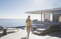Женщина в купальнике ходит по солнечному современному роскошному внутреннему дворику с видом на океан — стоковое фото