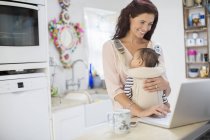 Mother holding baby boy and using laptop in domestic kitchen — Stock Photo