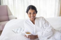 Portrait smiling woman in bathrobe drinking tea on bed — Stock Photo