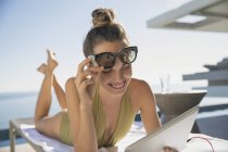 Усміхнена жінка в купальнику і сонцезахисних окулярах за допомогою цифрового планшета, купається на кріслі для відпочинку на сонячному патіо — стокове фото