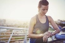 Female runner resting checking smart watch fitness tracker on sunny urban footbridge — Stock Photo