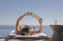 Женщина загорает, используя цифровой планшет на шезлонге на солнечном патио с видом на океан — стоковое фото